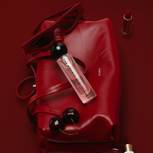 Cherry 60ml perfume bottle by The Bombay Duo on red leather bag with cherries, sunglasses, and lipstick. A sensual fragrance in a bold, stylish setting.